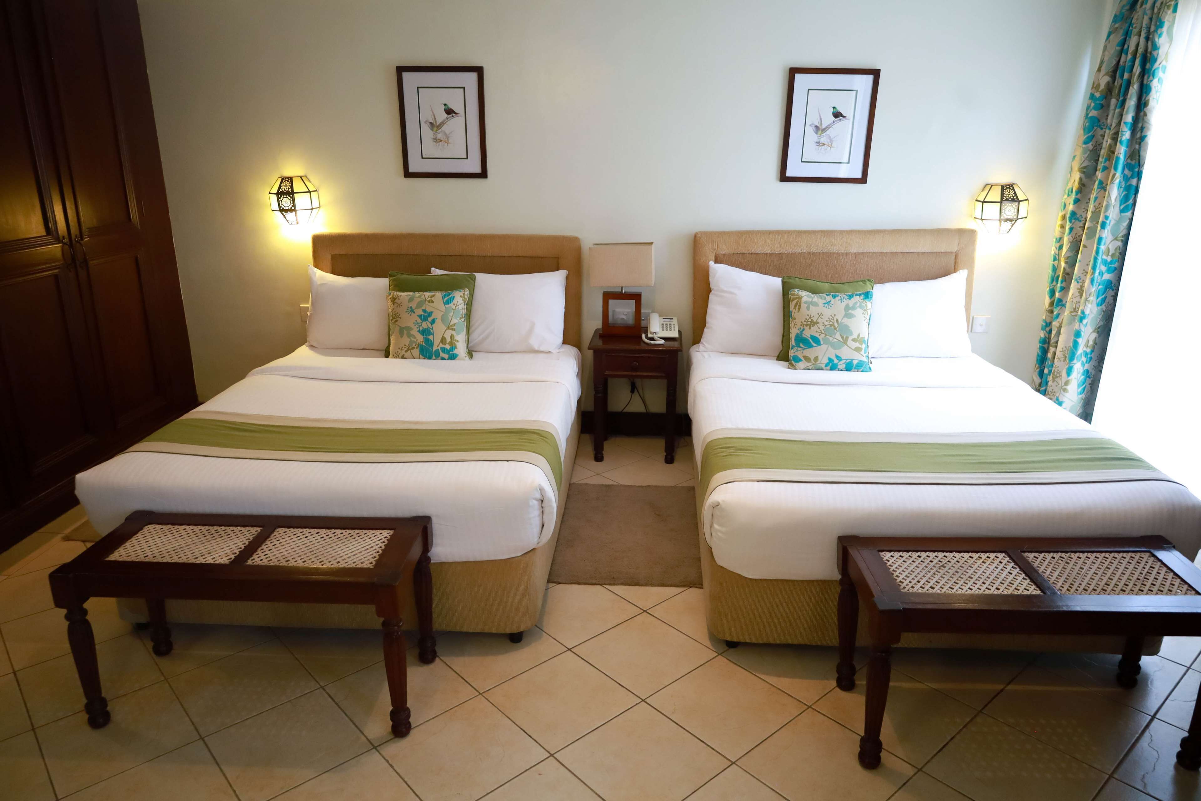 A hotel room with two beds, each with a headboard, a bedside table, and a lamp. There are framed pictures on the wall above the beds.