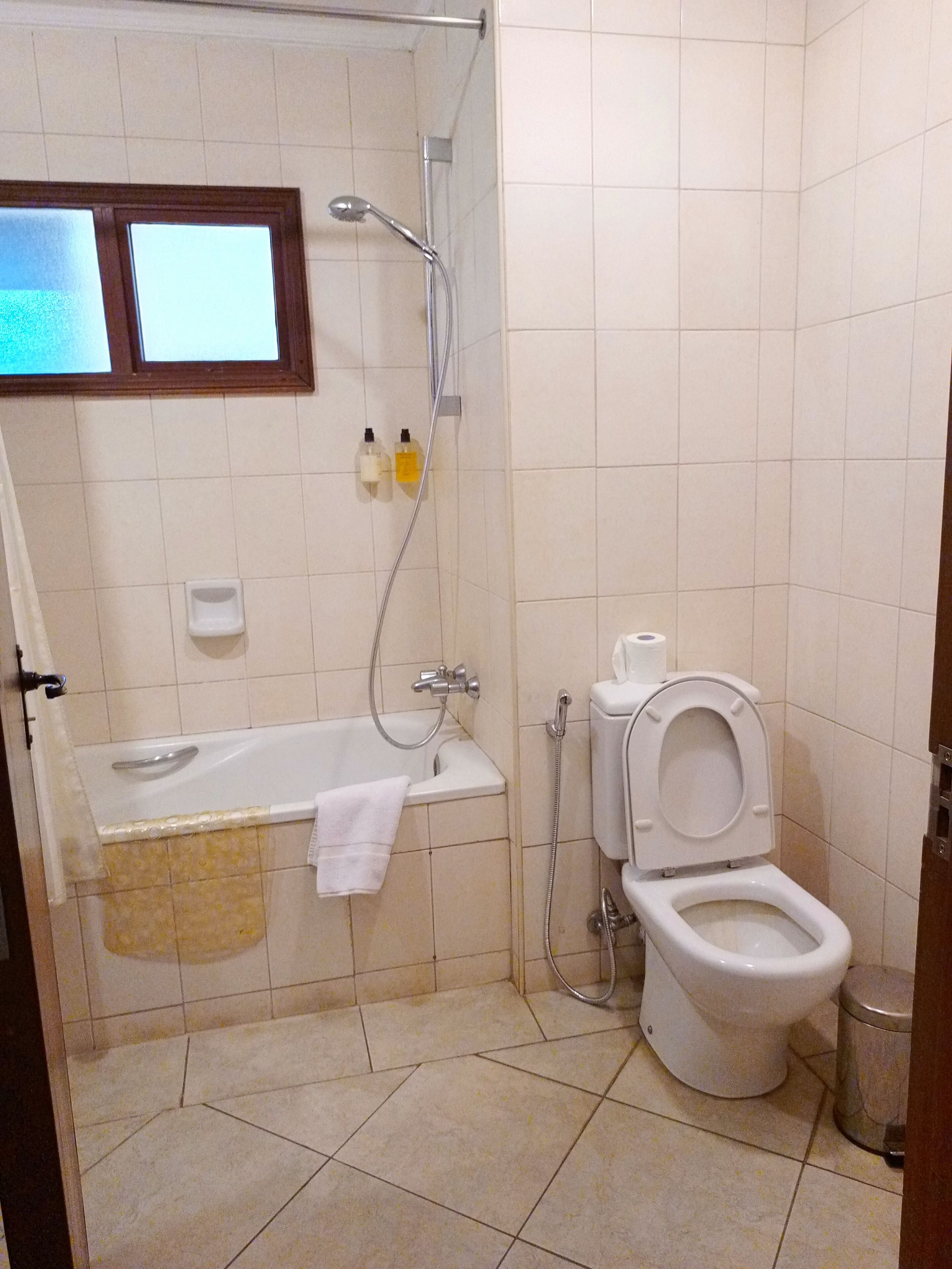 A small bathroom with a bathtub, toilet, and shower. There is a window above the bathtub and a towel hanging on the wall.