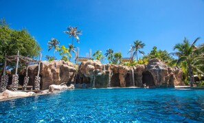 Una piscina azul cristalina con una cascada artificial de rocas, rodeada de palmeras y un entorno tropical.