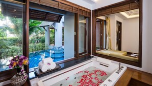 A luxurious bathroom with a large bathtub, a glass-enclosed window overlooking a pool, and a mirror reflecting the room. - Royal Muang Samui Villas (Koh Samui)