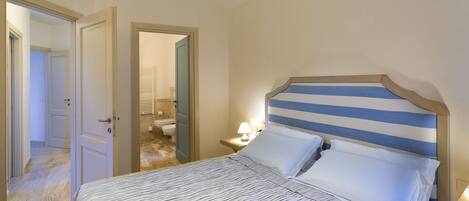 Ein Schlafzimmer mit gestreifter Kopfteil, einem Bett mit Streifenmuster, einem Nachttisch mit Lampe und einem durchgehenden Türspalt sichtbaren Badezimmer.