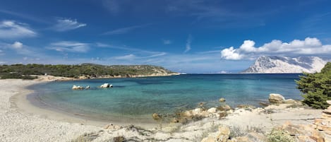 A coastal landscape with clear turquoise waters, rocky shorelines, and distant mountains under a blue sky.
