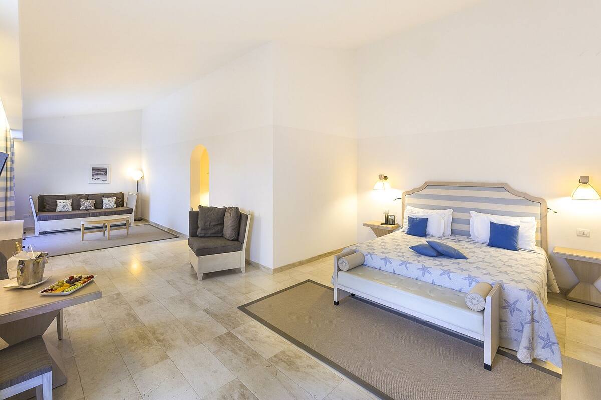 Een hotelkamer met een bed, een sofa, een kleine tafel met een schaal fruit en een nachtkastje met een lamp.