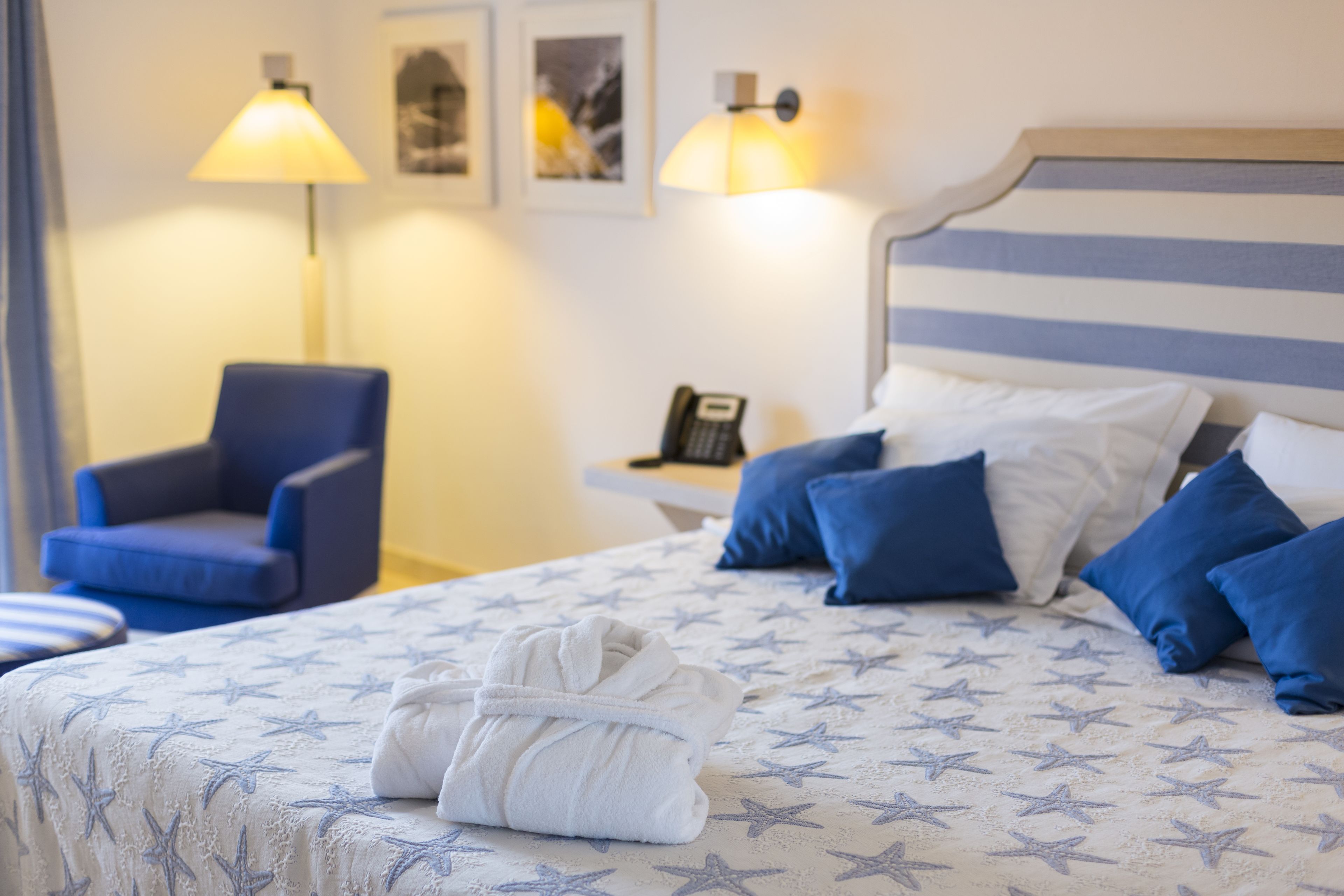 Una cama bien hecha con una colcha de estampado de estrellas, una bata doblada sobre la cama, un sillón azul y una mesita de noche con una lámpara y un teléfono.