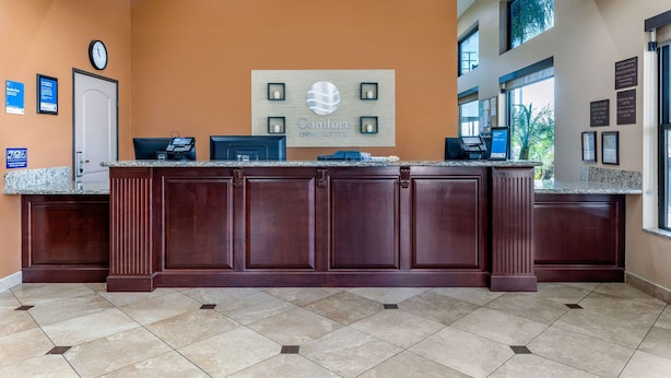 A reception desk with a clock, a sign for Comfort Inn & Suites, and a window with a view of palm trees. - Comfort Inn & Suites San Bernardino (Colton)