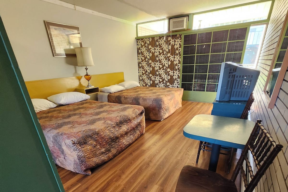 A room with two beds, a table, a chair, a lamp, and an air conditioning unit.