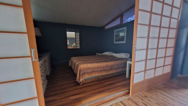 A traditional Japanese-style room with a wooden floor, a bed with a patterned bedspread, a small bedside table, and a mirror on the wall. - Tiki Village (Vernon)