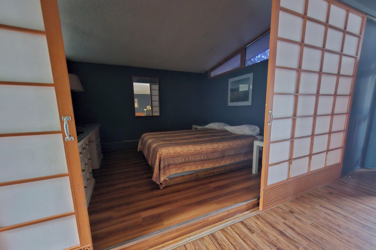 A traditional Japanese-style room with a wooden floor, a bed with a patterned bedspread, a small bedside table, and a mirror on the wall.