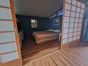 A traditional Japanese-style room with a wooden floor, a bed with a patterned bedspread, a small bedside table, and a mirror on the wall. - Tiki Village (Vernon)