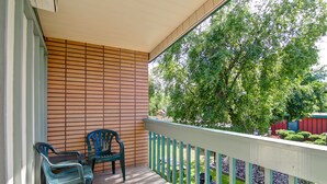 A balcony with a wooden floor, two green plastic chairs, and a view of a garden with trees and a red building in the background. - Tiki Village (Vernon)