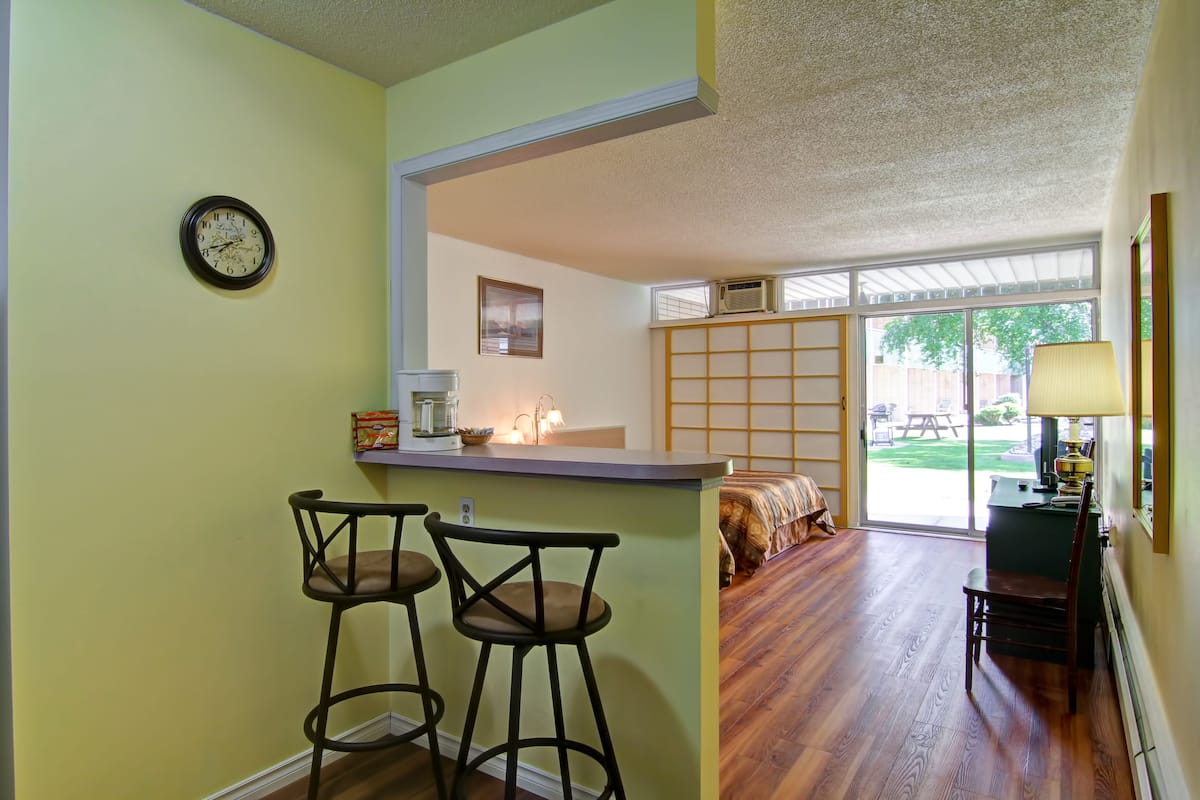 A kitchen with a bar area, two stools, a clock, and a view of a dining area with a table and chairs.