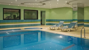 Una piscina interior con agua cristalina, rodeada de tumbonas y una pared de azulejos con una decoración en tonos verde y azul.