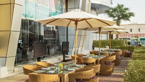 Un espacio exterior para comer con sillas y mesas de mimbre, sombrillas y un edificio moderno al fondo.