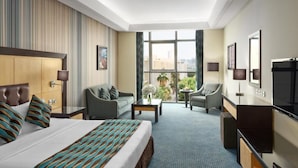 Habitación de hotel con una cama grande, un sofá, un escritorio y una televisión. Se ve una ventana con vistas a la ciudad.