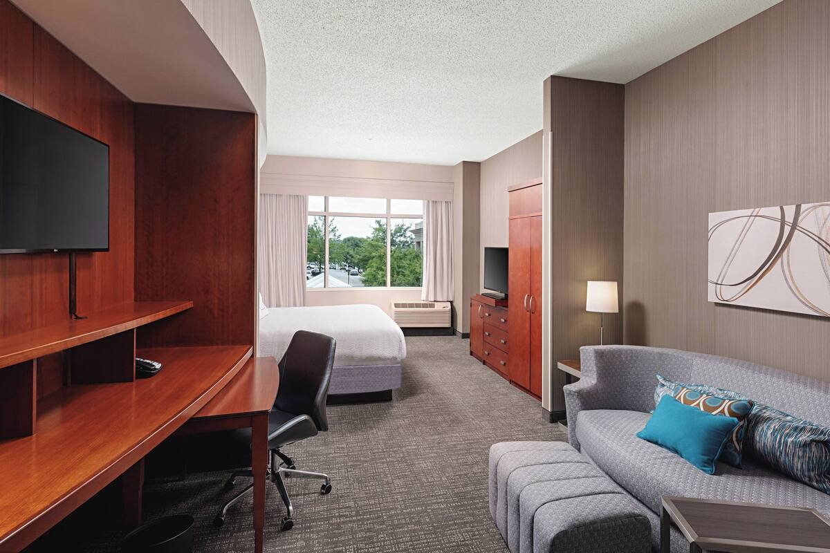 A hotel room with a bed, a desk, a TV, a sofa, and a window with a view of trees.