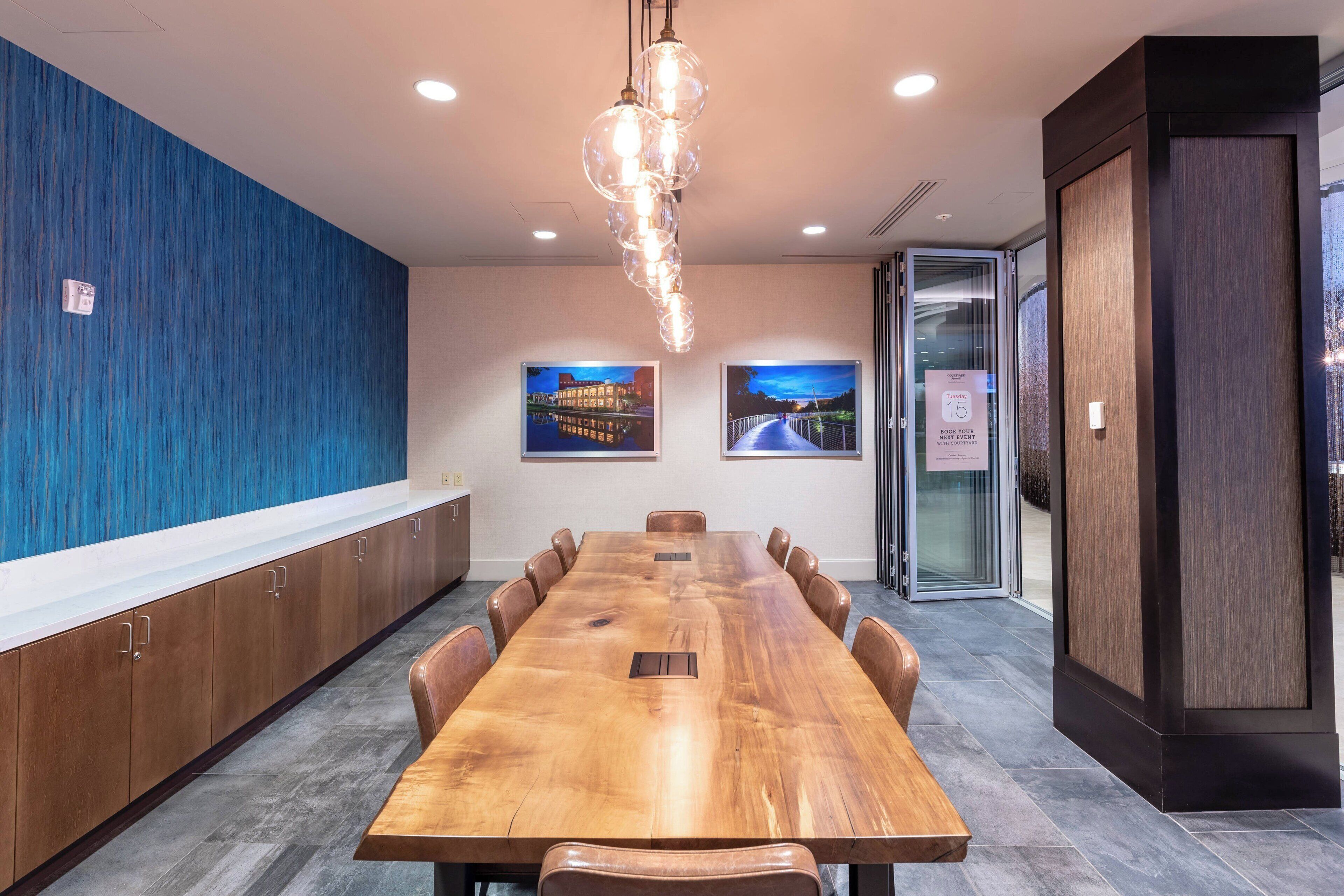 A conference room with a long wooden table, brown chairs, a hanging light fixture, and two framed pictures on the wall.