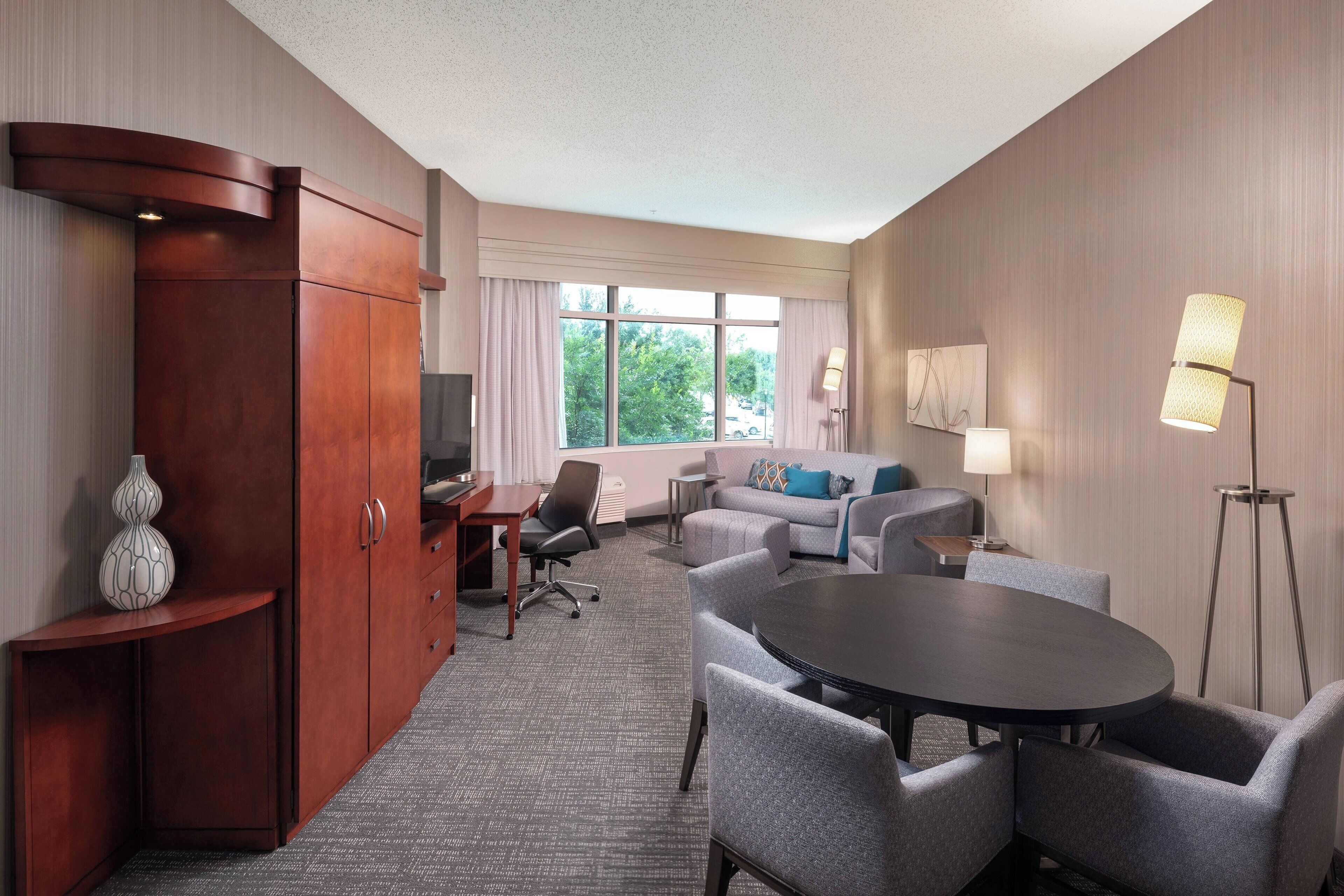 A hotel room with a wooden cabinet, a dining table with chairs, a sofa, and a lamp.