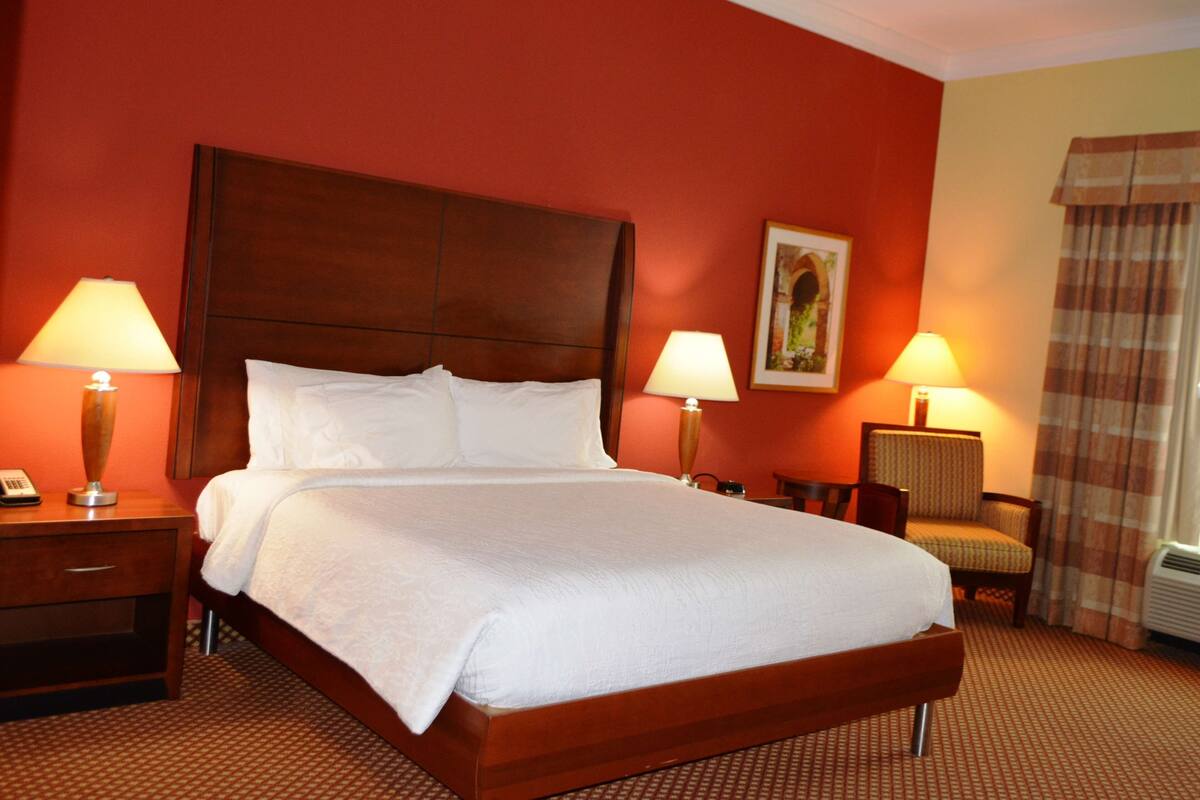 A hotel room with a large bed, two bedside lamps, a chair, a painting on the wall, and a curtain.