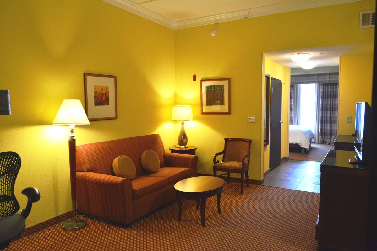 A hotel room with a yellow wall, a sofa, a chair, a small table, and a lamp. There is a bed in the background.