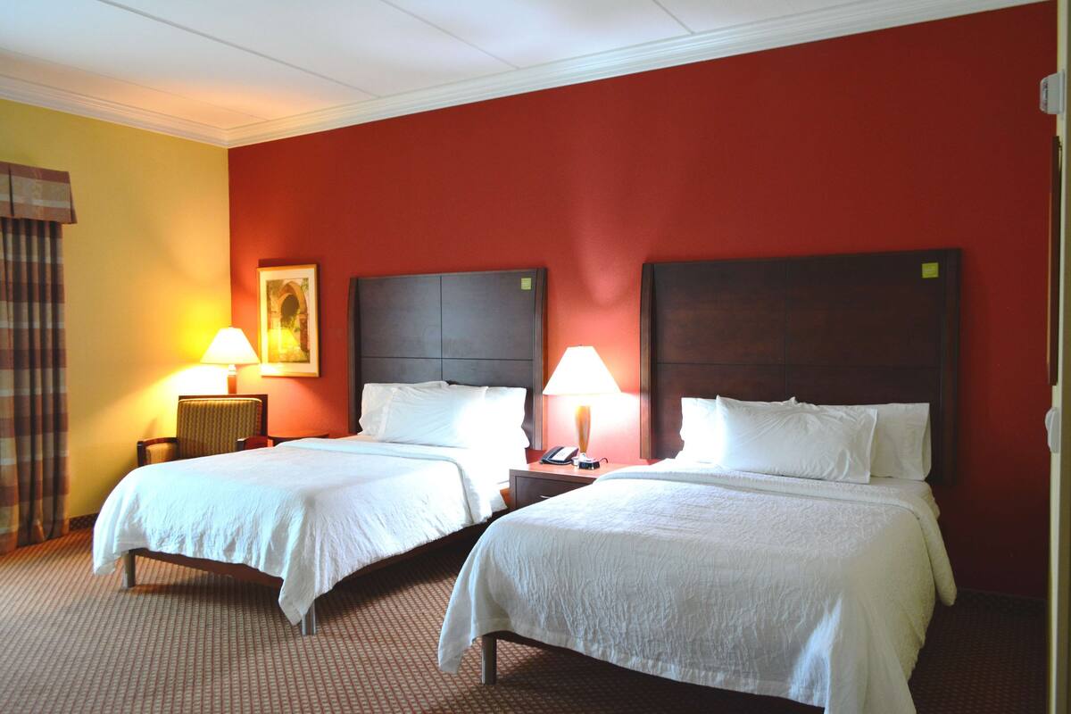 A hotel room with two beds, each with white linens, a headboard, bedside tables with lamps, and a framed picture on the wall.