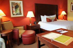A hotel room with a bed, a nightstand, a desk, a chair, and a red ottoman.