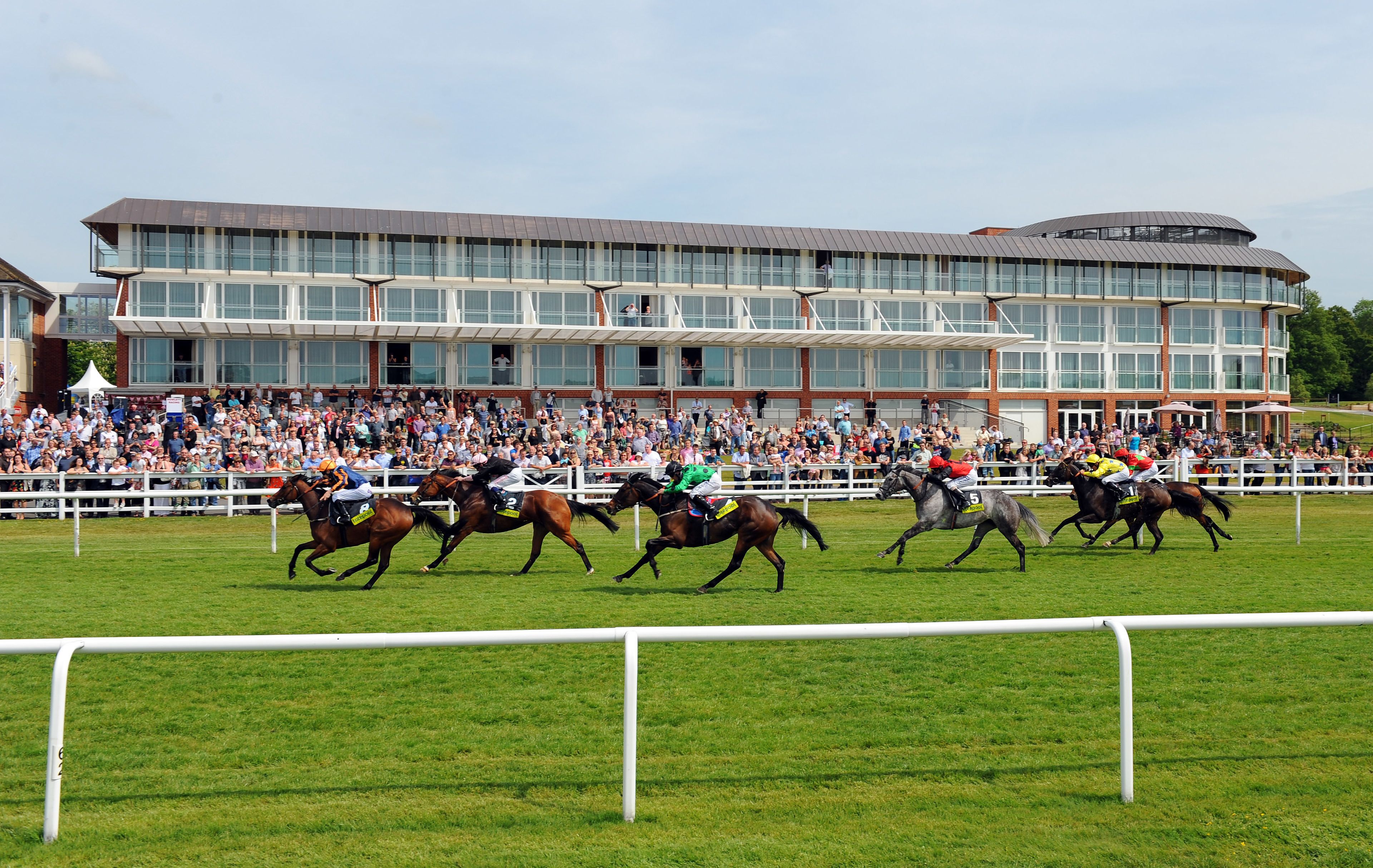 A horse race with jockeys on horses, a crowd of spectators, and a large building in the background.