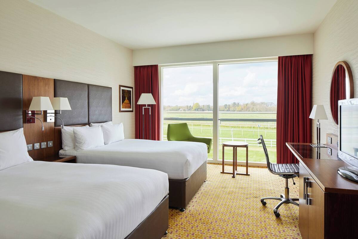 A hotel room with two beds, a desk, a chair, and a view of a tennis court.