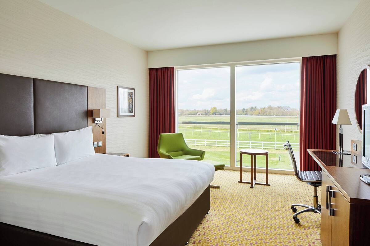 A hotel room with a large bed, a desk with a computer, a green chair, and a view of a field through the window.