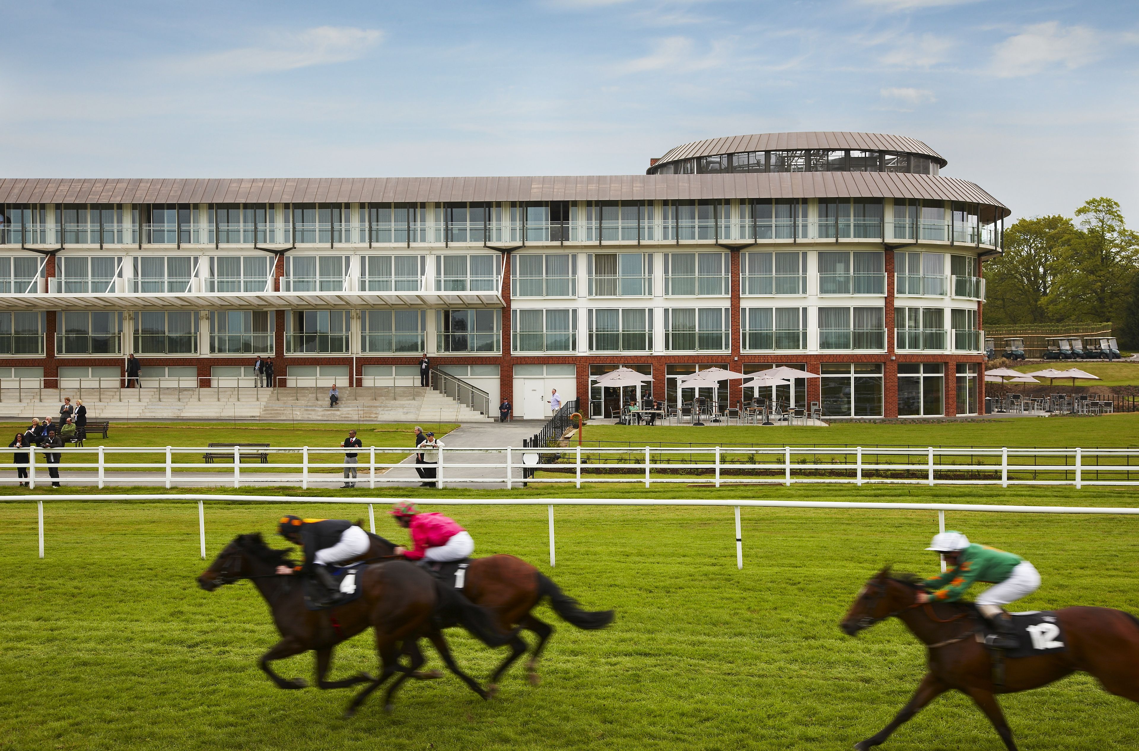 A multi-story building with a large glass facade, a circular section on the top, and a fenced area with a few people and horses.