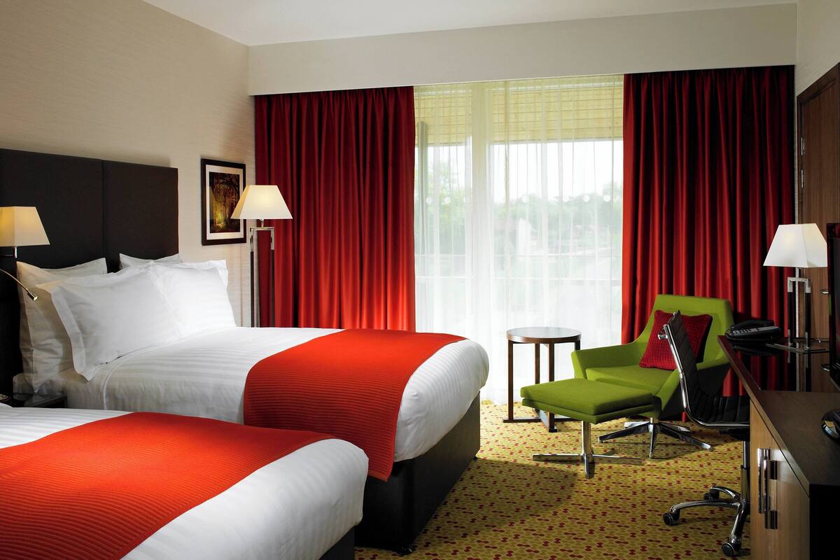 A hotel room with two beds, a green armchair, a desk, and a window with red curtains.