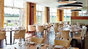 Un restaurante espacioso con numerosas mesas y sillas, amplias ventanas que ofrecen vistas al exterior, y modernos focos circulares en el techo.