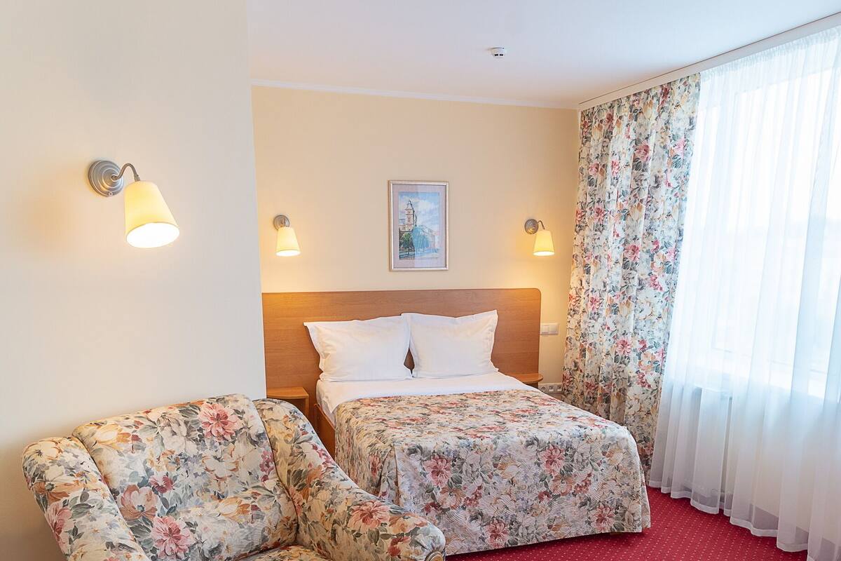 Quarto de hotel com cama, duas poltronas florais, tapete vermelho e janela com cortinas leves.