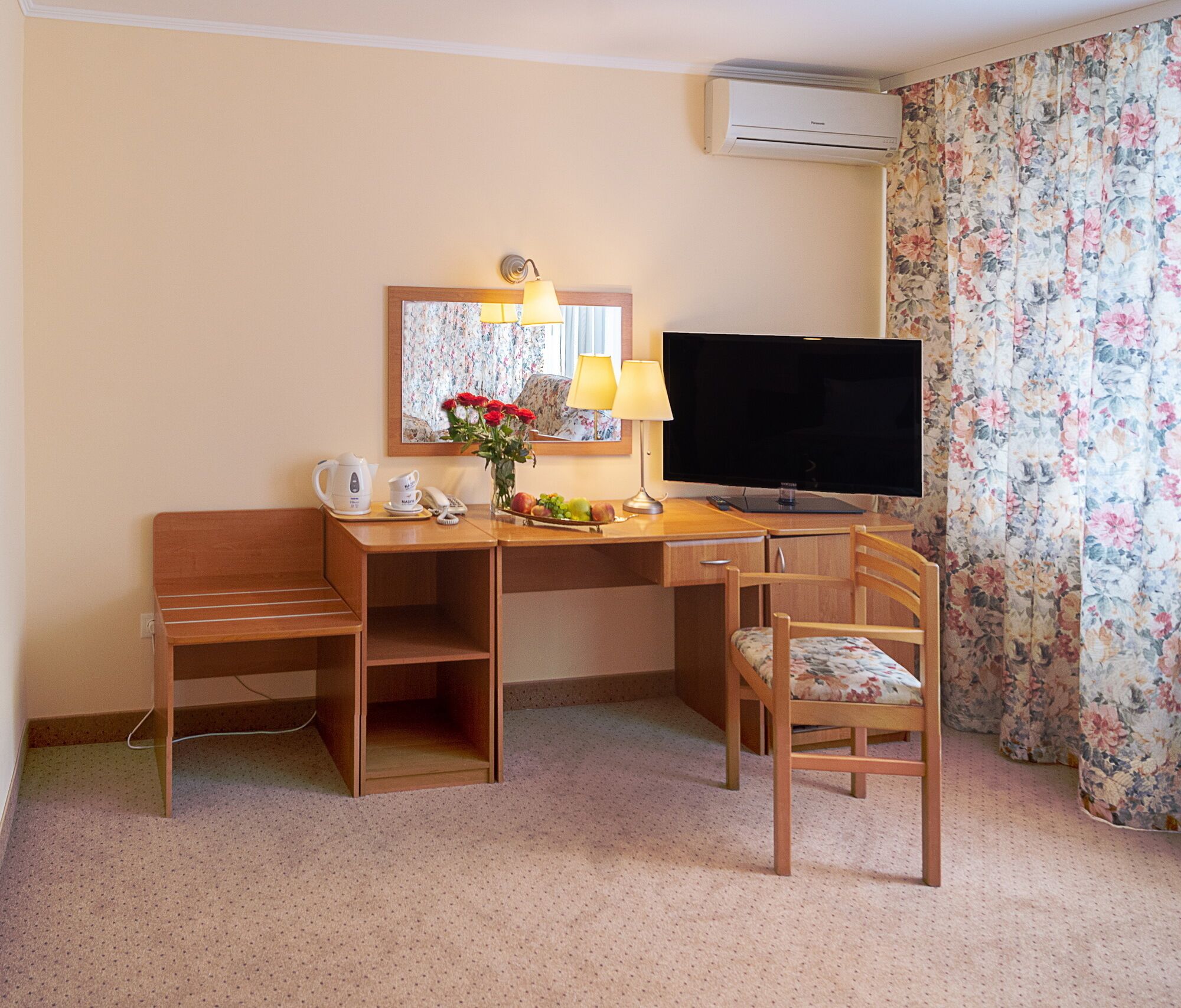 Habitación de hotel con un escritorio de madera, televisor, silla, espejo y cortina estampada con motivos florales.