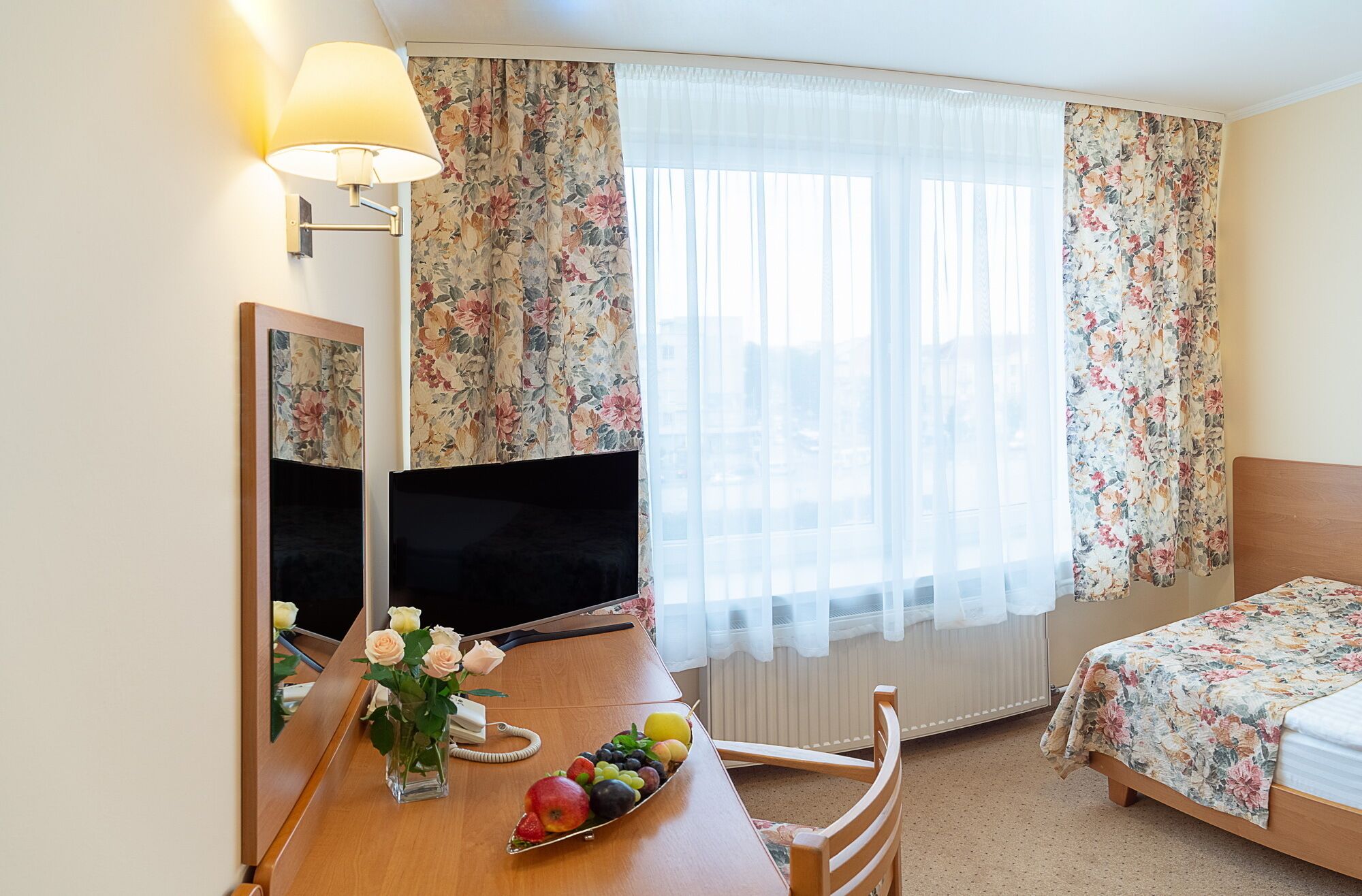 A hotel room with a bed, a desk with a TV, a mirror, and a window with floral curtains.