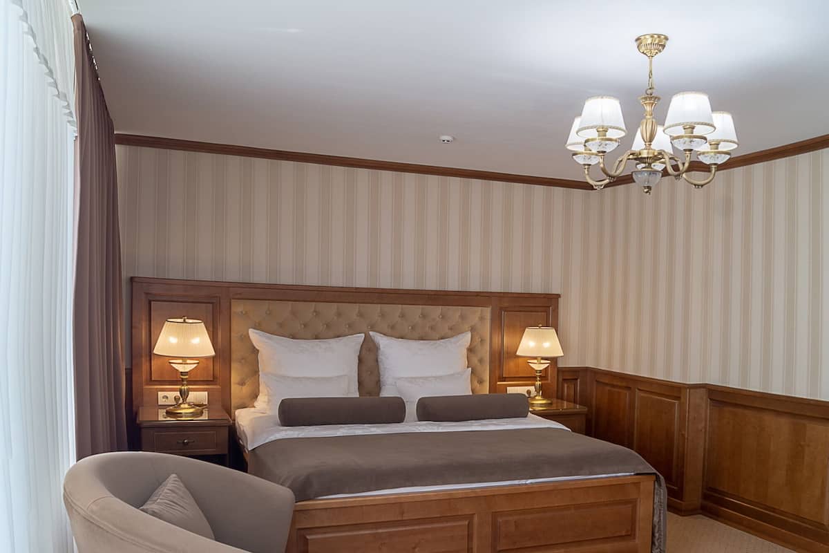 Quarto de hotel com uma cama grande, duas luminárias de cabeceira, uma poltrona e um candelabro.
