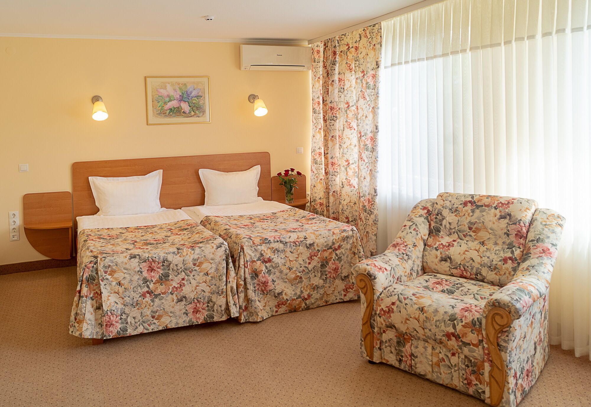A hotel room with a bed, two bedside tables, a chair, a floral curtain, and a wall-mounted air conditioner.