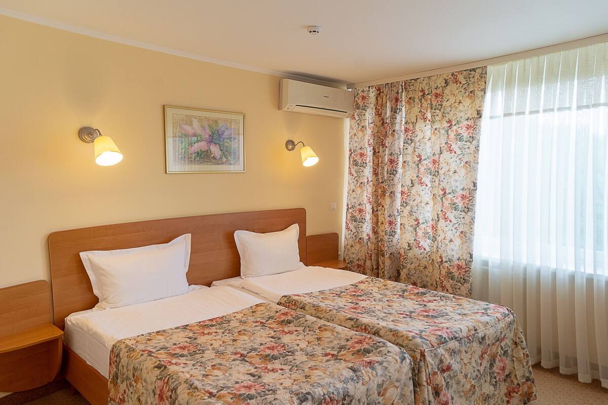 Quarto duplo com duas camas, cabeceira de madeira, colcha floral, um quadro, ar condicionado fixado na parede e uma janela com cortinas leves.
