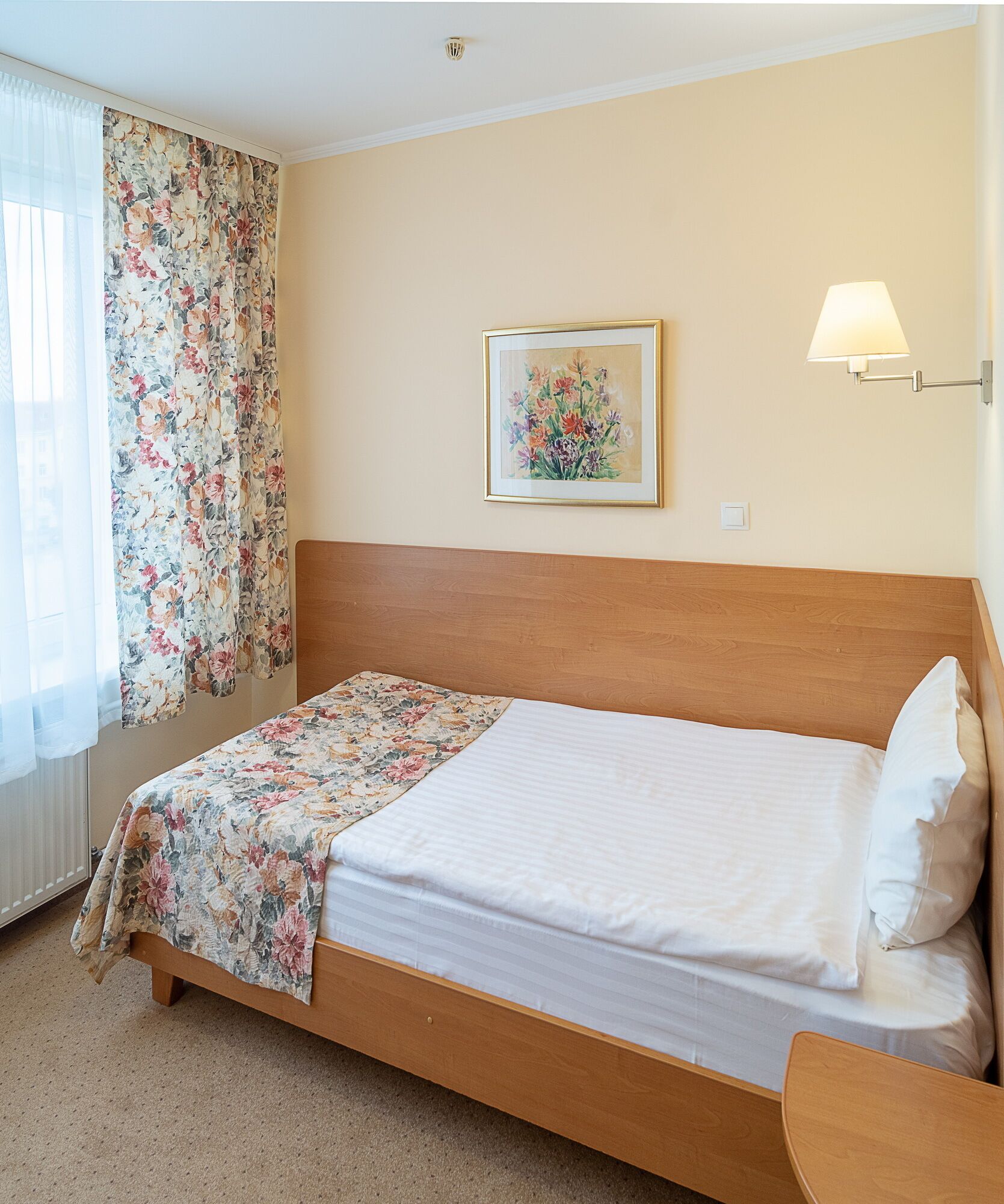 Cama bem arrumada com colcha florida, quadro floral emoldurado na parede e abajur.