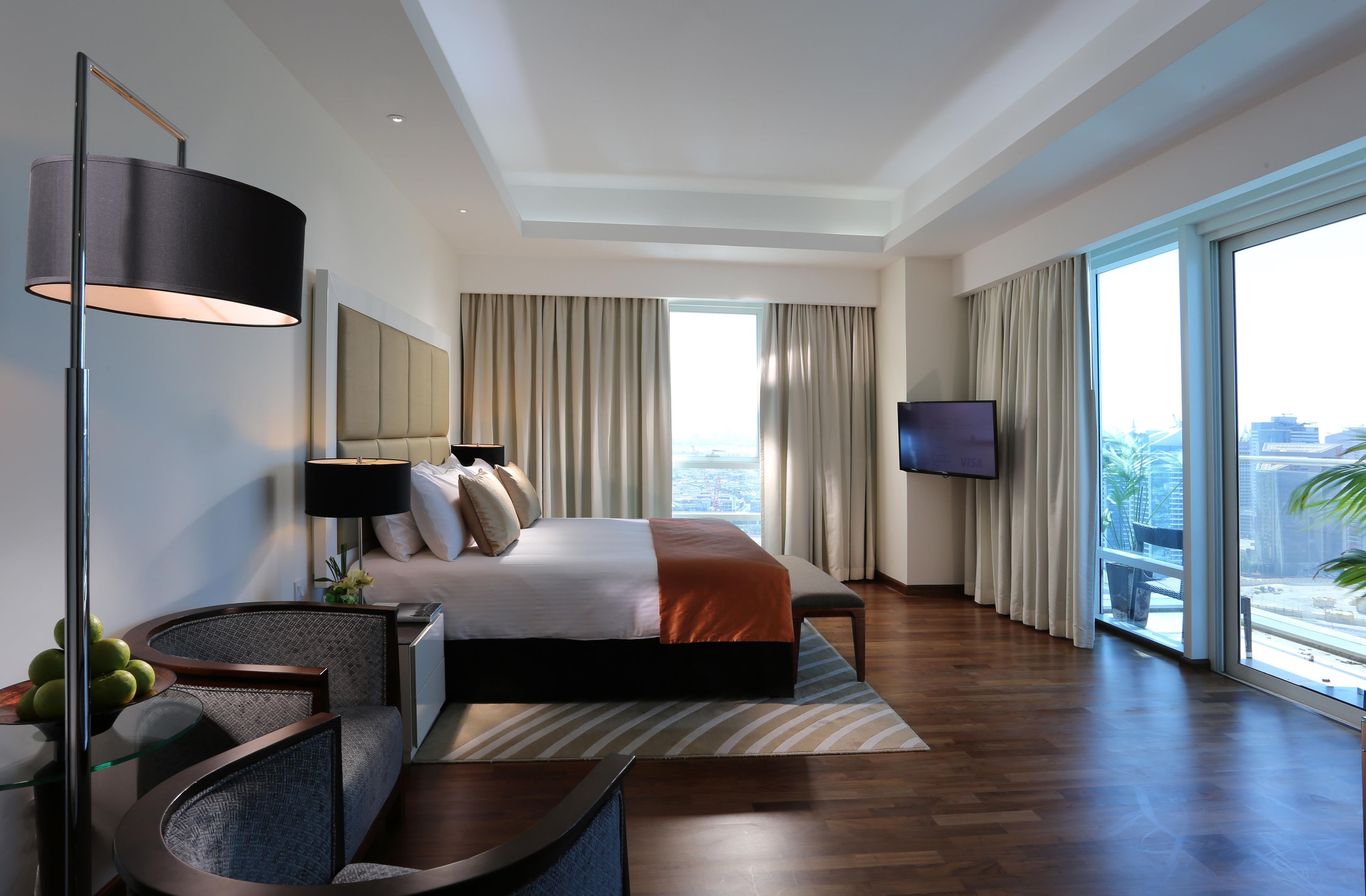 A modern hotel room with a large bed, a flat-screen TV, a desk with a lamp, and a view of the city through large windows.