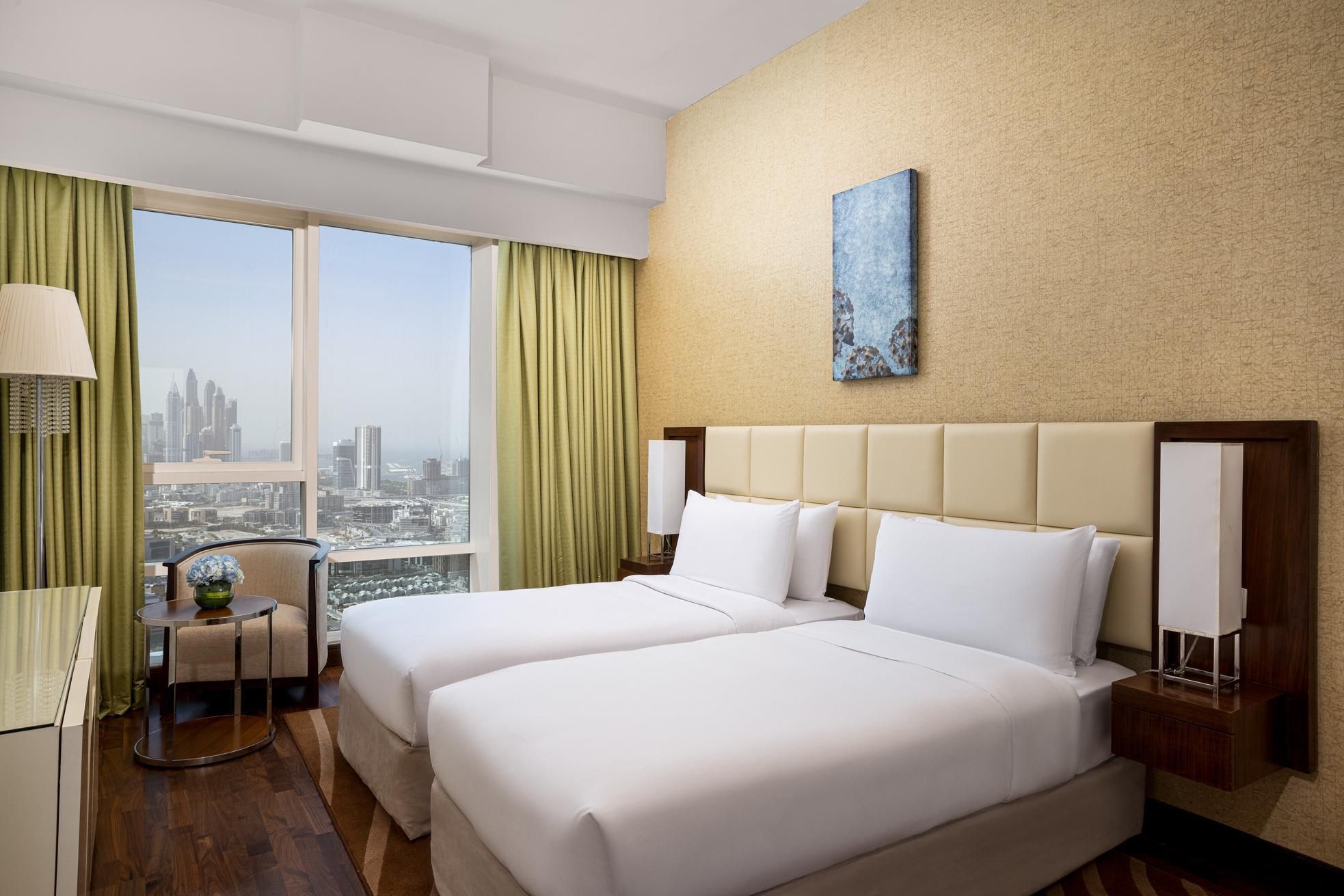 Habitación de hotel con dos camas, vista a la ciudad, un cuadro en la pared y una mesita con una lámpara.