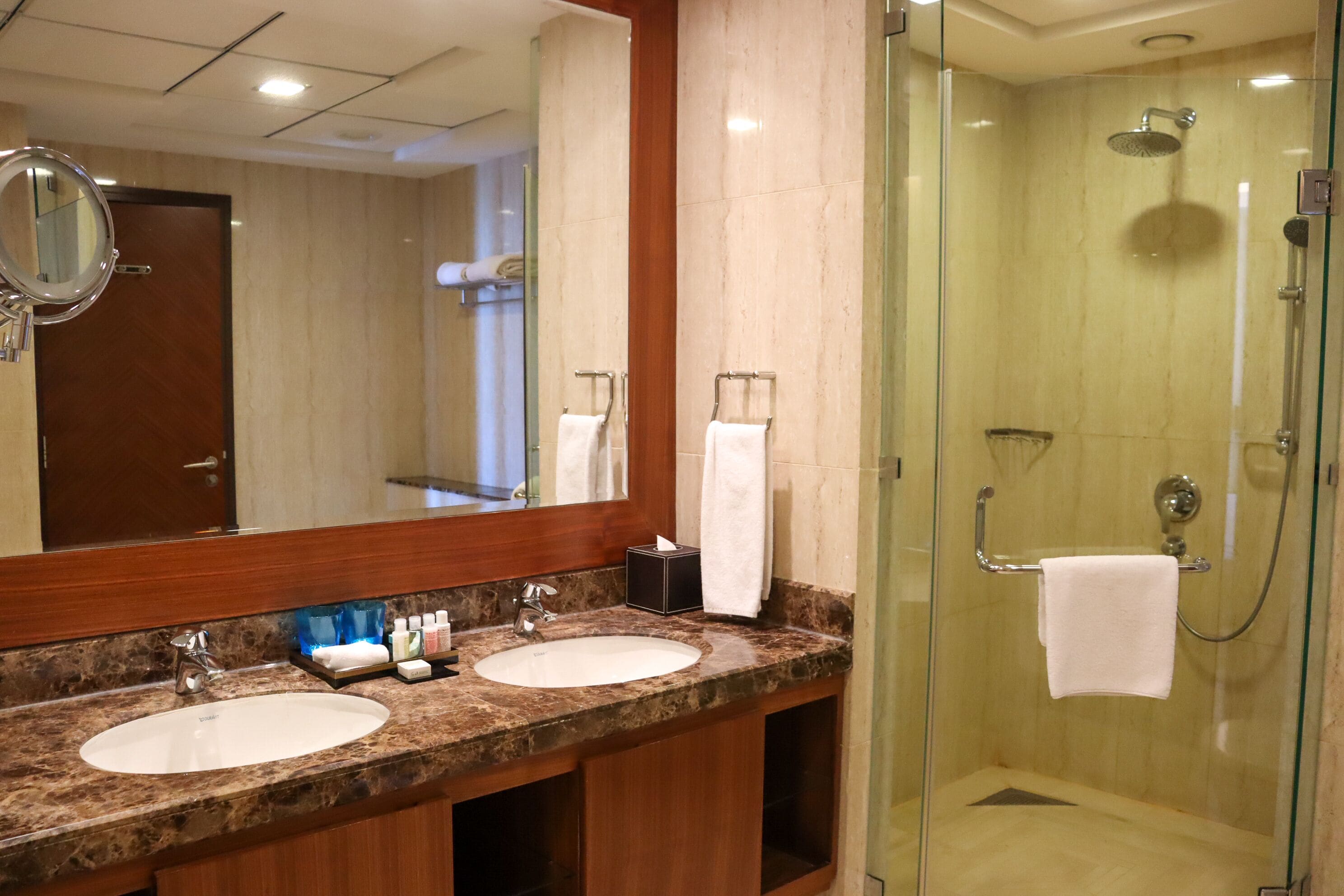 A hotel bathroom with two sinks, a large mirror, and a glass-enclosed shower area.