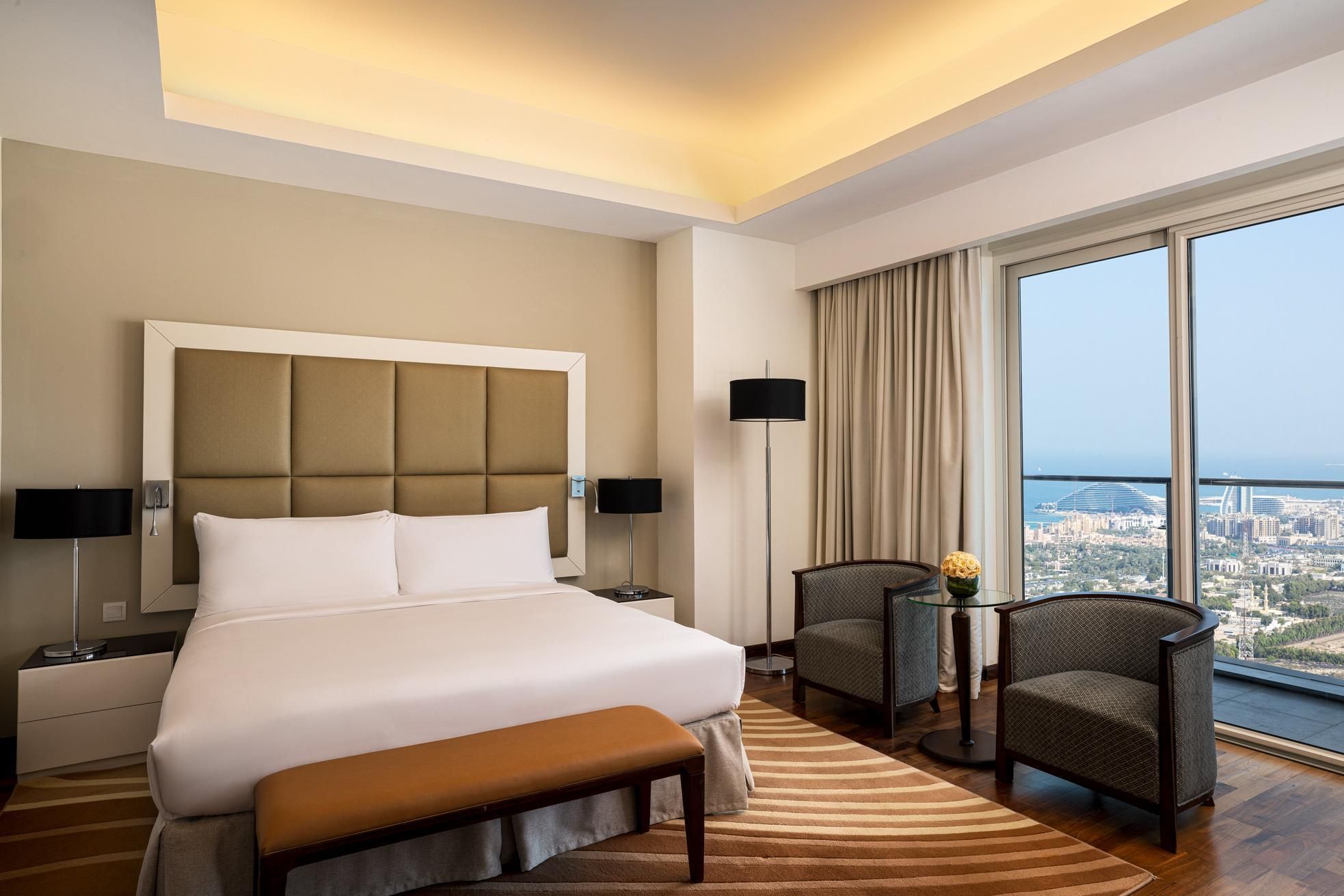 Ein modernes Hotelzimmer mit einem großen Bett, zwei Sesseln, einem kleinen Tisch und Blick auf die Stadt.