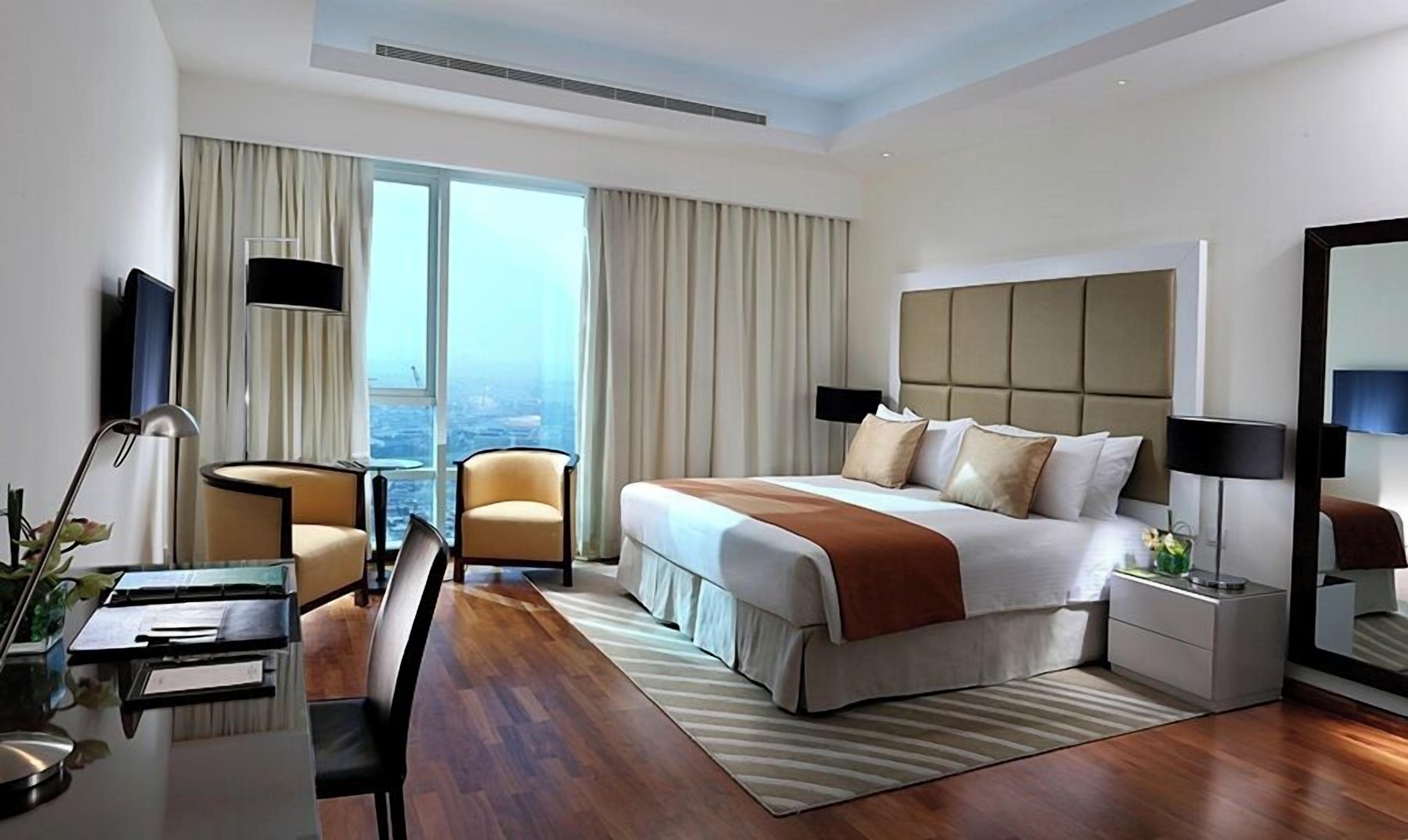 A hotel room with a bed, a desk, a chair, a TV, and a view of the city.