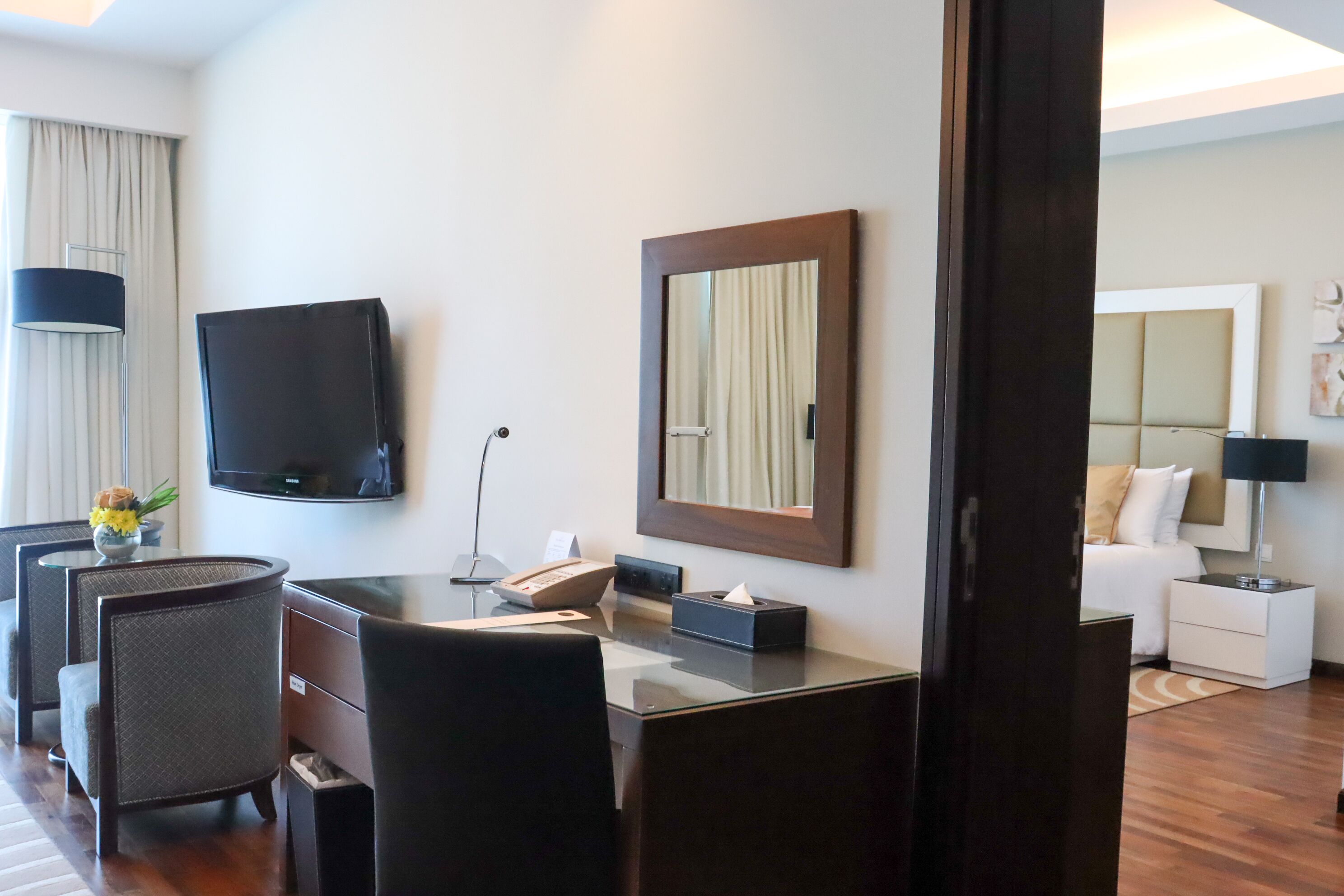 A hotel room with a desk, chairs, a television, a mirror, a bed, and a nightstand.