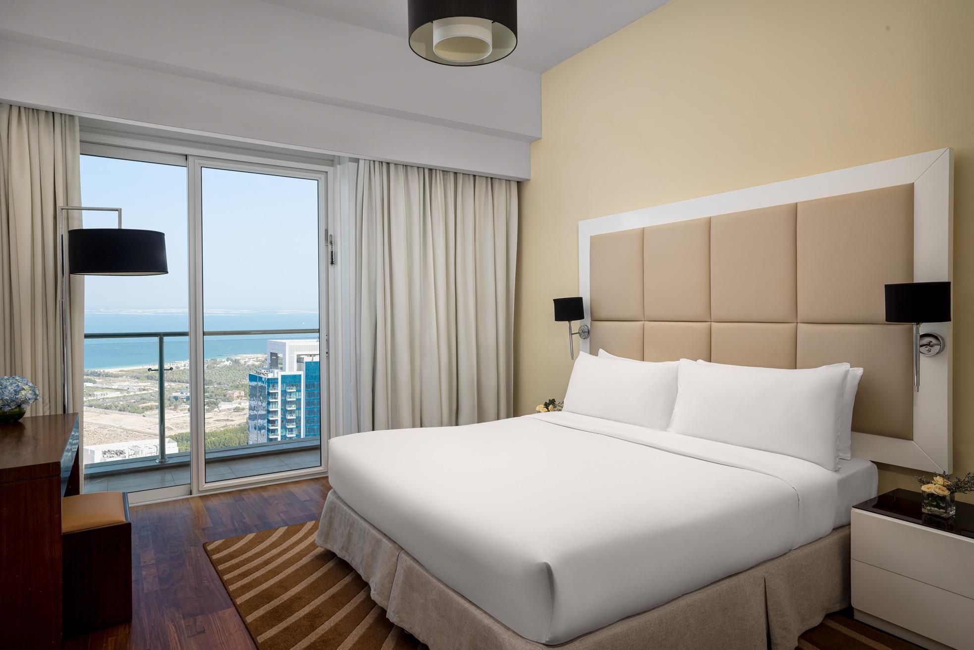 Habitación de hotel con una cama grande, una mesita de noche, un escritorio y vistas al mar y edificios a través de la ventana.