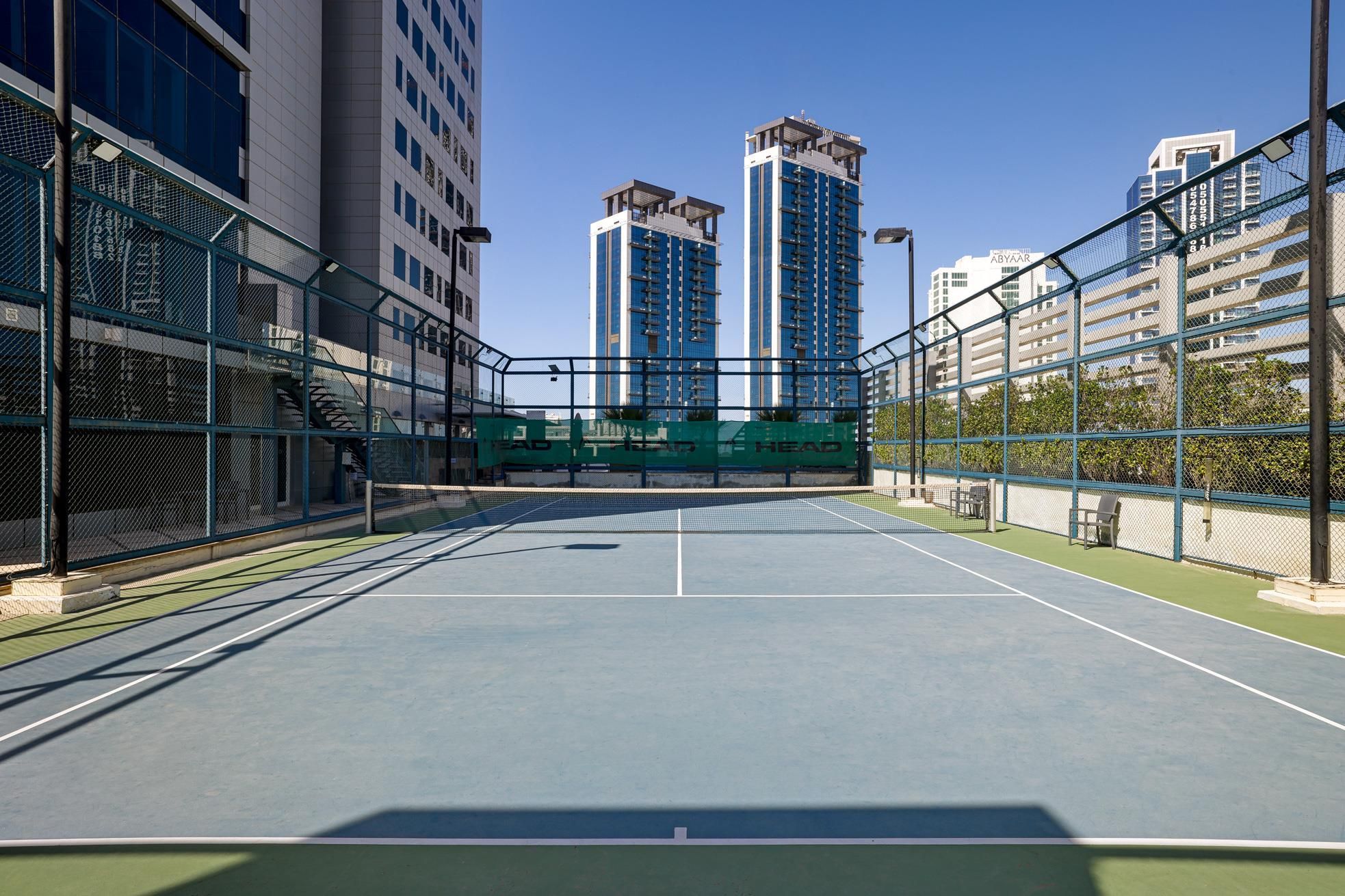 An outdoor tennis court with a net, surrounded by high-rise buildings.
