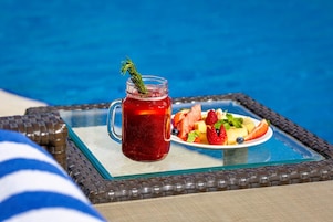 Un jarro de vidrio con una bebida roja y un plato de fruta sobre una mesa junto a una piscina.