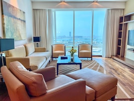 A modern living room with a sofa, two armchairs, a coffee table, and a TV. Large windows offer a view of the cityscape.