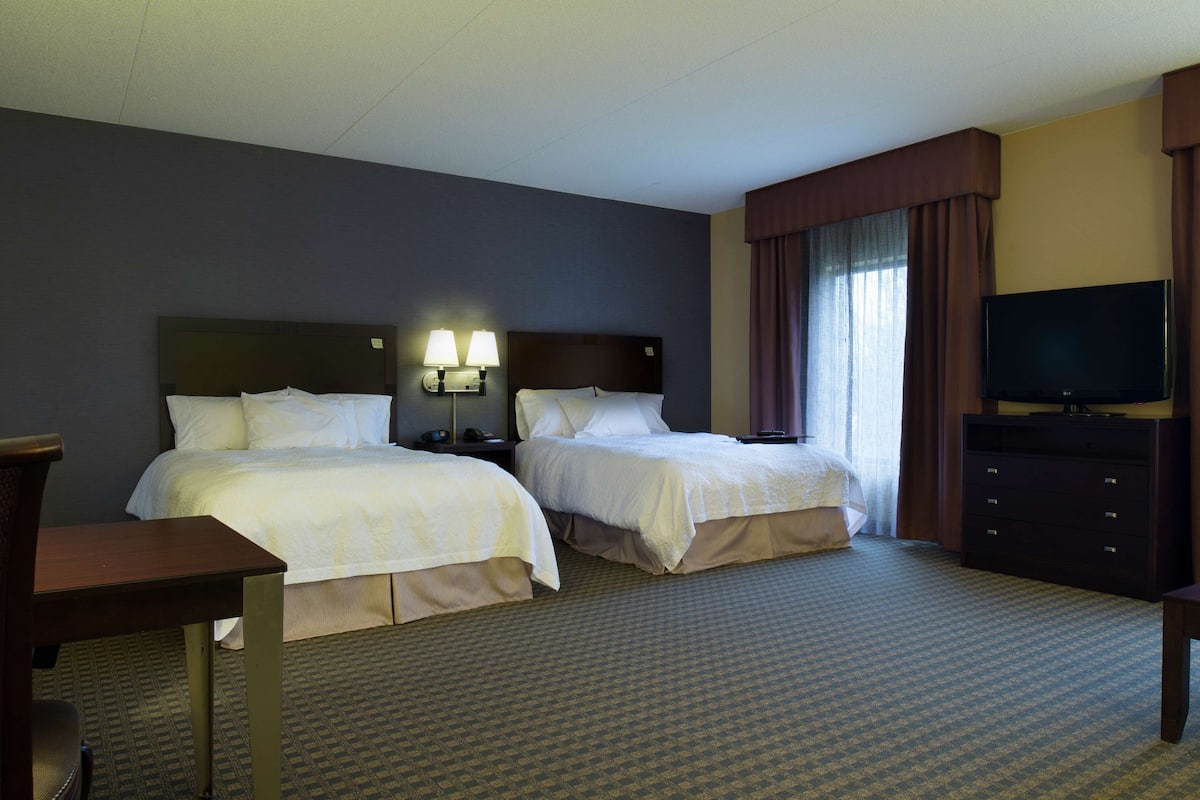 A hotel room with two beds, a desk, a dresser, a television, and a window with curtains.