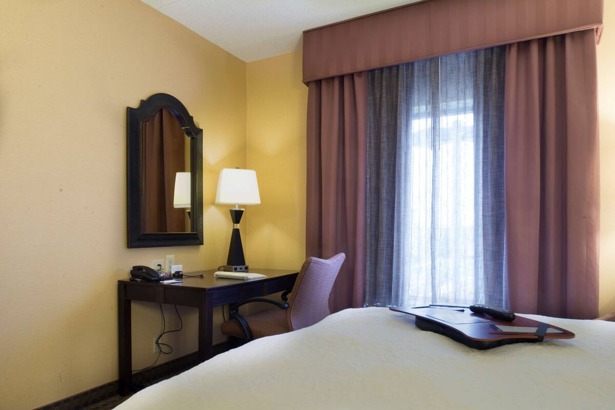 A hotel room with a bed, a desk, a chair, a lamp, a telephone, and a window with curtains.